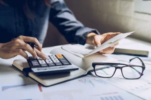 Person using a calculator and reviewing documents at a desk with papers, a notebook, eyeglasses, and information on workers compensation benefits.