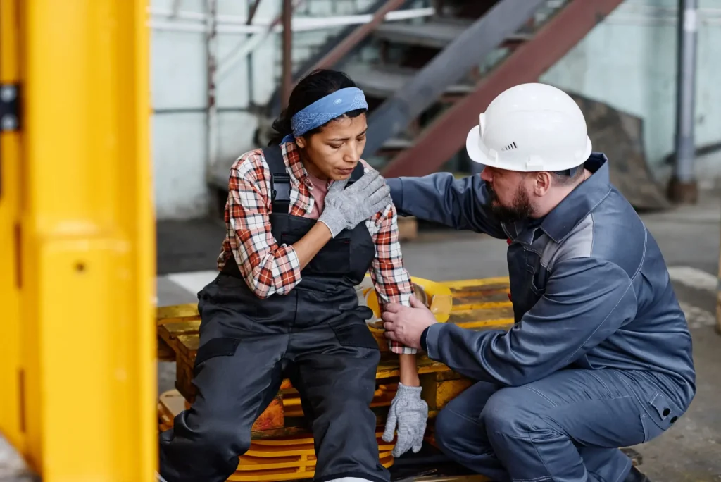 Injured factory worker receiving assistance from a supervisor after a workplace accident in an industrial setting.