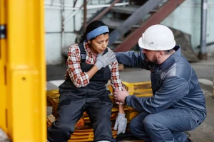 A worker in overalls sits holding her shoulder while another worker in safety gear, who recently passed a drug test, kneels beside her offering assistance in an industrial setting.