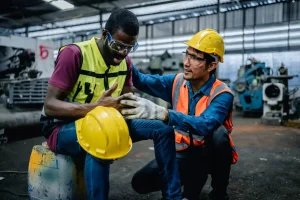 Two workers in safety gear are in a factory; following a workplace accident, one appears injured and is sitting, while the other kneels beside him, offering support.