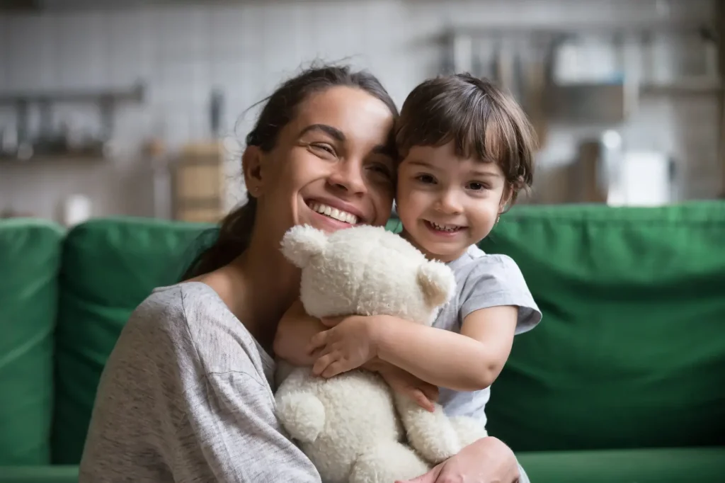 A parent and a young child sit on a green couch, smiling and hugging a white teddy bear in a cozy indoor setting.