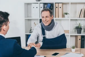 Smiling man with a neck brace and arm sling, appearing to be an independent contractor, shakes hands with another person across a desk in an office setting.