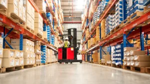 A warehouse worker operates a red forklift between tall shelves stacked with boxes and packages on pallets.
