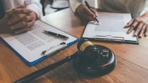 Two people sit at a table with legal documents, a pen, and a clipboard, next to a judge's gavel and two wedding rings on a wooden block.