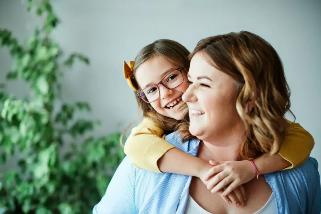 A smiling young girl with glasses and a yellow bow hugs a woman from behind; both are looking happy. A green plant is blurred in the background.
