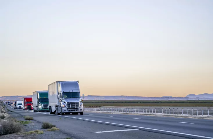A line of semi-trucks driving on a road during sunset, with mountains visible in the background, mirrors the journey of Monroe Workers Compensation Attorneys as they navigate complex legal terrain.