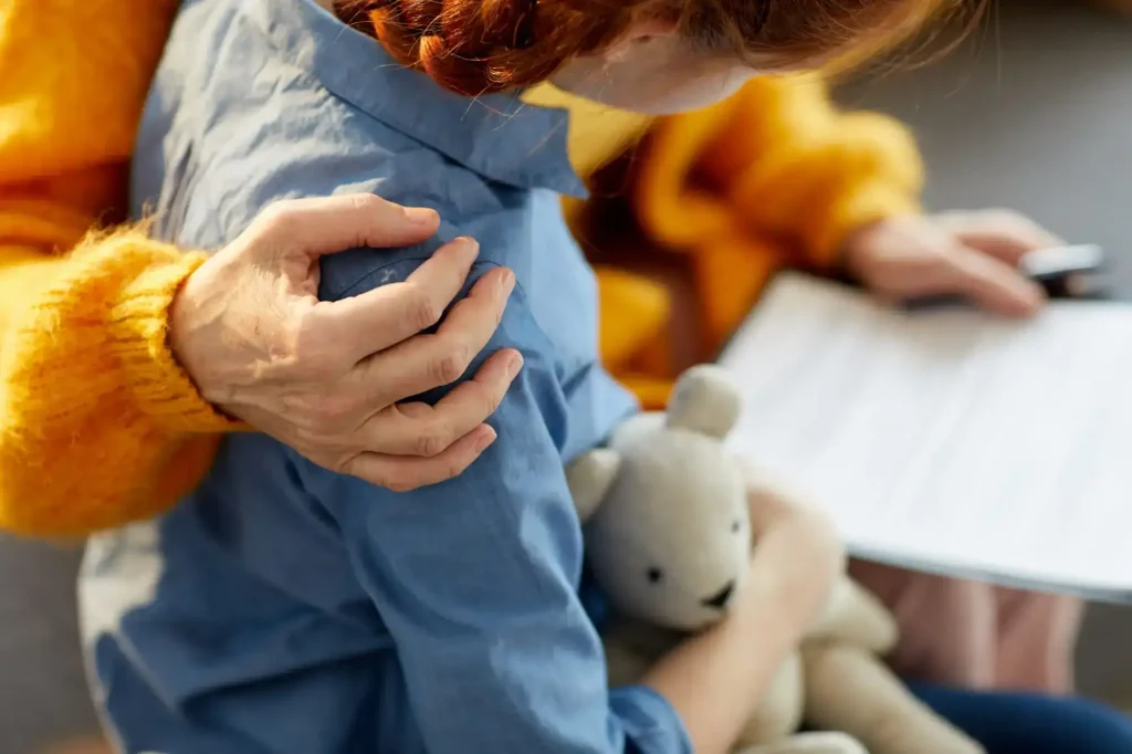 An adult comforts a child holding a teddy bear while showing them a document or paper.