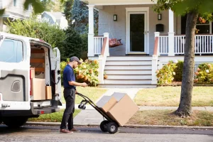 A delivery person moves cardboard boxes on a hand truck from a van parked in front of a suburban house.
