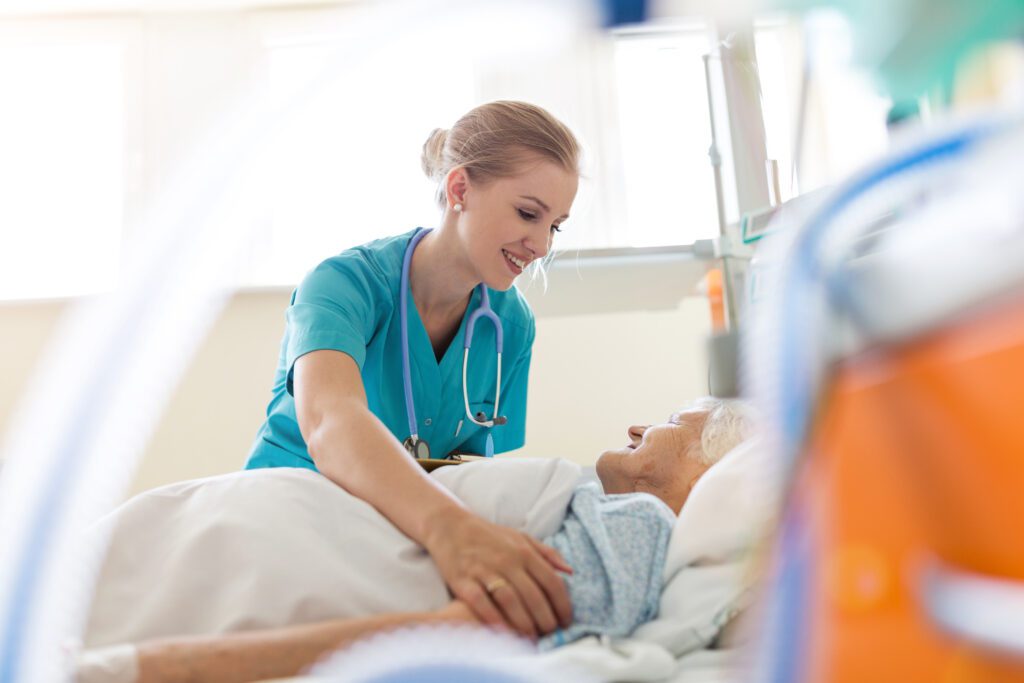 A nurse in blue scrubs with a stethoscope around her neck is smiling and attending to an elderly patient lying in a hospital bed, embodying the same compassion you'd expect from Charlotte Family Lawyers.