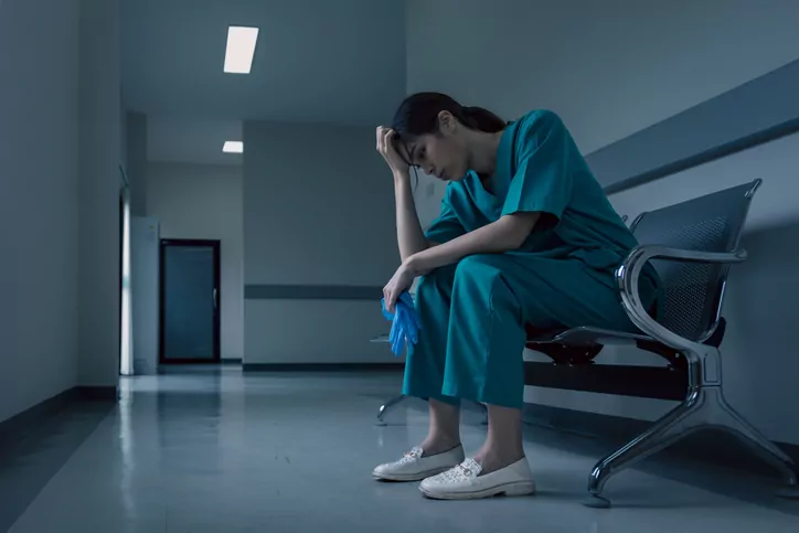 A person in medical scrubs sits on a bench in a hallway, holding a pair of gloves and resting their head on their hand, appearing fatigued—a scene all too common for those needing the services of a Charlotte Workers Compensation Lawyer.