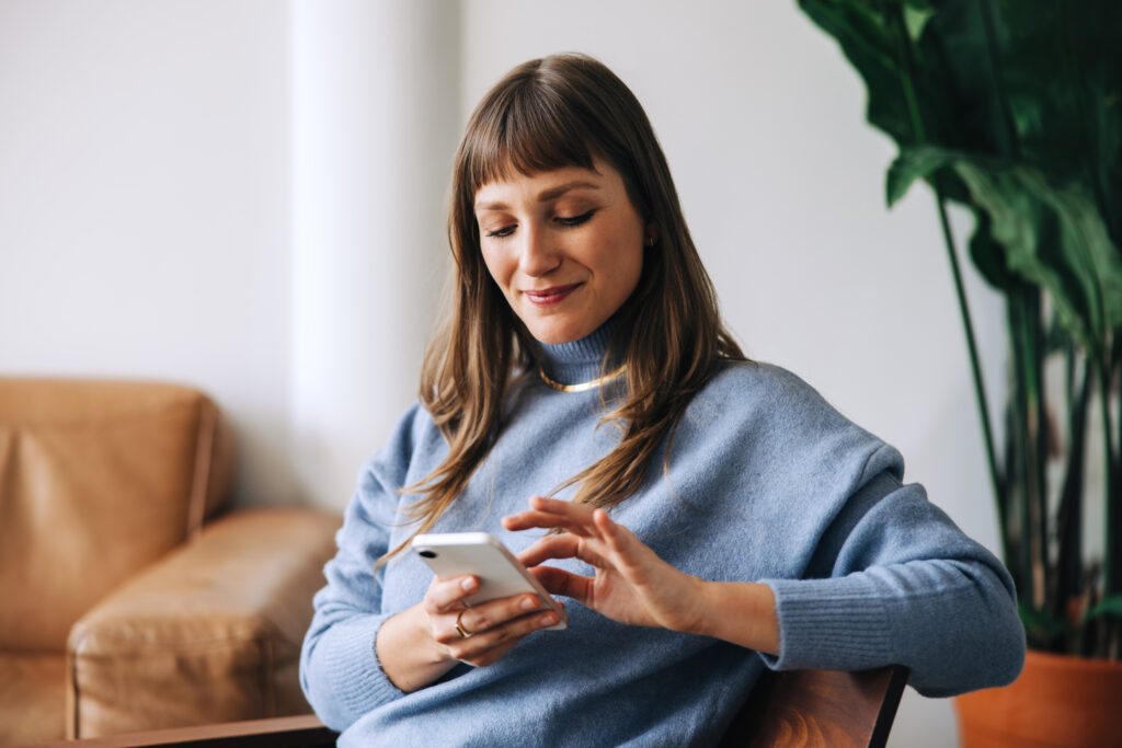 A woman sitting in a chair, wearing a blue sweater, is smiling and using a smartphone. In the background of her cozy living room with a large plant and beige sofa, she might be researching the best Gastonia Divorce Lawyer for her needs.