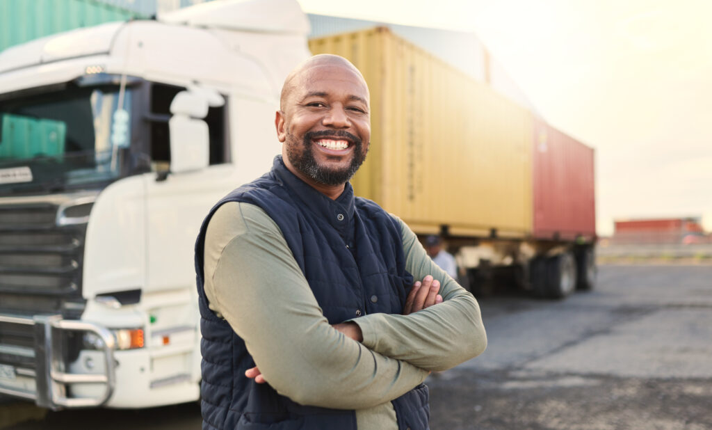 A smiling man stands with arms crossed in front of a large truck with shipping containers at a lot, showing the resilience and determination one might find in a Charlotte Workers Compensation Lawyer.