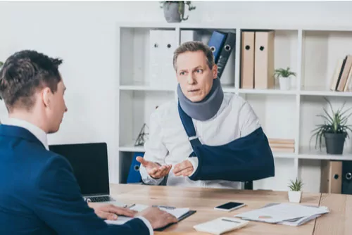 Two men in a meeting, with one man wearing a neck brace and arm sling, gestures while talking. They are seated at a desk with paperwork and office supplies present, alongside shelves in the background.