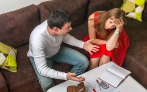 A man and woman sit on a couch, engaged in a serious conversation. The man gestures with his hands while the woman looks stressed, resting her head in one hand. Open notebooks and papers are on the table, suggesting they might need advice from a Monroe Workers Compensation Attorney.