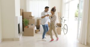 A couple dances in a living room surrounded by moving boxes and a bike near a large window, their joyous moment undisturbed despite the recent visit from Charlotte Family Lawyers.