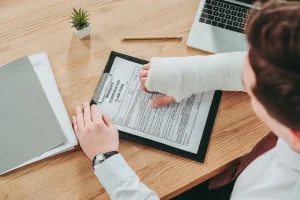 A person with a bandaged arm fills out a workers' compensation claim form at a desk with a laptop, pencil, and folder.