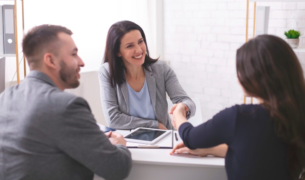 Three people in business attire sit at a table. One woman smiles while shaking hands with another woman, as a man watches and smiles. Tablets and papers are on the table, possibly discussing legal matters handled by Charlotte Family Lawyers.