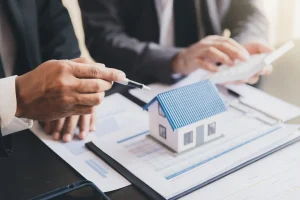 Two people in business attire discuss documents with a model house, pen, and calculator on the table, representing a real estate or mortgage meeting.