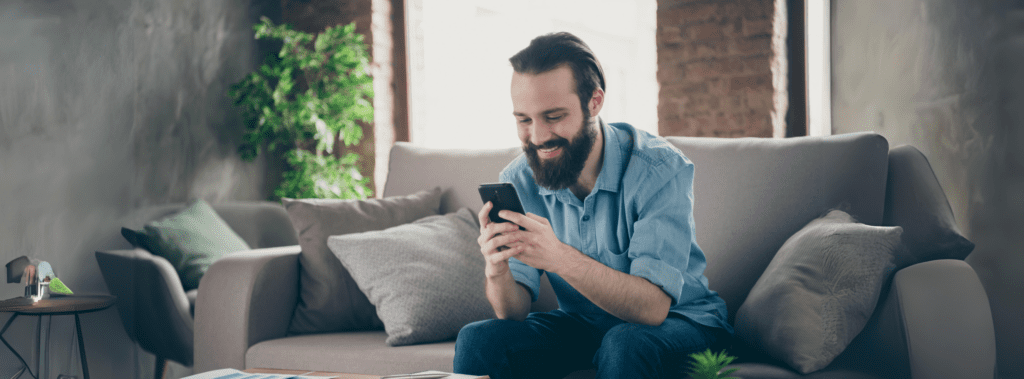 A man with a beard sits on a couch in a living room, smiling while looking at his phone.