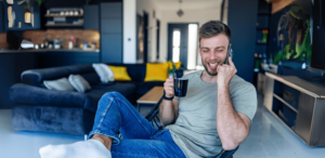 A man sits comfortably in a modern living room, holding a mug and talking on a smartphone. He is casually dressed and smiling, perhaps discussing some details.