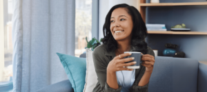 A woman sits on a couch, smiling and holding a cup of coffee. She appears relaxed in a cozy living room setting, perhaps taking a well-deserved break from her busy day as one of the leading Charlotte Family Lawyers.