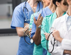 A group of medical professionals wearing scrubs and lab coats and holding stethoscopes stand in a line.