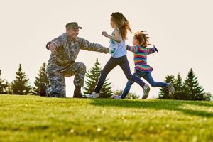 A person in military uniform kneels on the grass with arms outstretched as two children run towards them in a park setting.