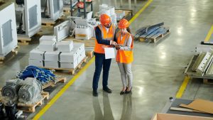 Two workers in orange safety vests and hard hats discuss a clipboard while standing in an organized, spacious warehouse with machinery and equipment on pallets.