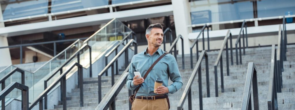 A man in a blue shirt and khaki pants stands on steps outside a modern building, holding a smartphone and a coffee cup, possibly waiting for his appointment with his Charlotte workers' comp lawyer about his weekly wage benefits.