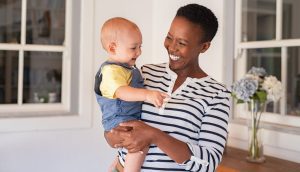A person holding a smiling baby in a well-lit room with white walls and large windows, reminiscent of a welcoming space curated by a Charlotte adoption lawyer. A vase with flowers can be seen in the background.