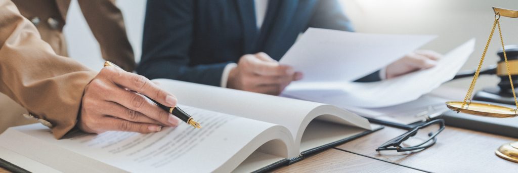 Two individuals are sitting at a desk, reviewing legal documents. One person is pointing at an open book with a pen, and a pair of glasses and a gavel are visible on the desk.