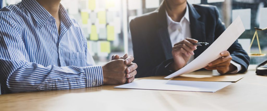Two people sit at a table reviewing a document. One person holds and points to the paper with a pen while the other listens attentively with hands clasped. A set of scales is visible in the background, hinting at the complexities of workers' compensation benefits in Charlotte.