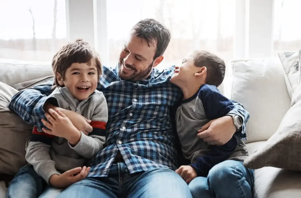 A man sits on a couch with two young boys, smiling and laughing together in a bright living room.