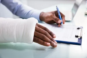 A person with a bandaged arm is filling out a form on a clipboard with a pen, next to an open laptop on a desk.