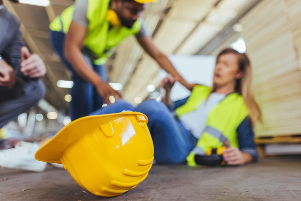 A yellow hard hat on the floor with a woman in a safety vest sitting and appearing injured, assisted by a coworker to show work injury and needing to consult with a Denver Workers Comp Lawyer.