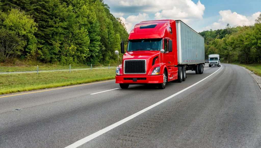 A red semi-truck with a trailer drives down a highway surrounded by trees, with another truck visible in the background—an everyday scene where a Charlotte Workers' Compensation Lawyer for Truck Drivers can help protect your rights after an accident.