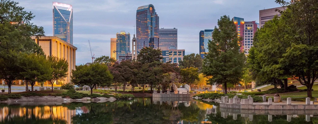 A cityscape view of Charlotte, North Carolina, with modern skyscrapers and buildings reflected in a park pond, surrounded by trees and greenery.