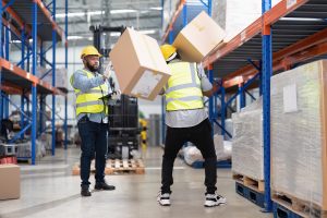 Two warehouse workers in safety vests and helmets handle cardboard boxes, with one box falling and getting hurt. consult with Charlotte Workers Comp Lawyer for help with case.