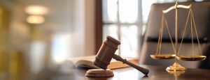 A wooden judge’s gavel rests on a desk beside an open book and a gold balance scale in a well-lit office.