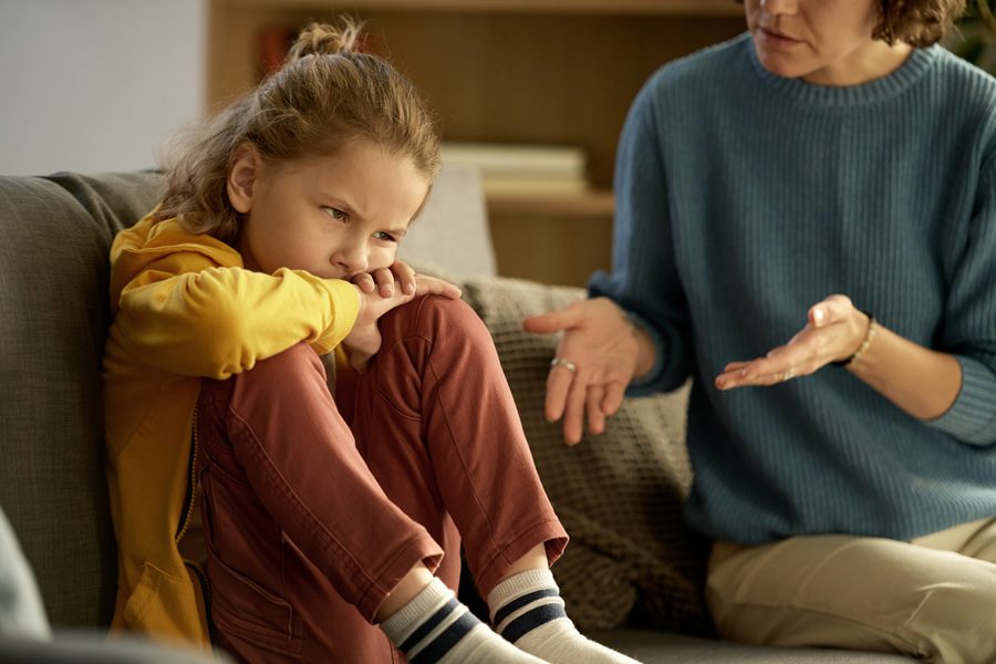 Pouting child sitting on couch with dissatisfied expression on his face while mother talking to him