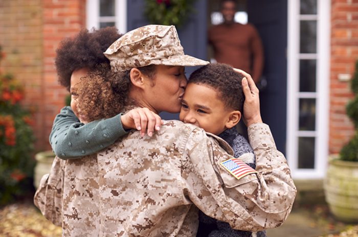 A female soldier is hugging her children outside of her home.