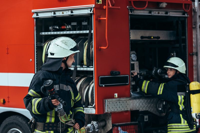 male firefighter in uniform holding water hose while colleague checking equipment in truck on street