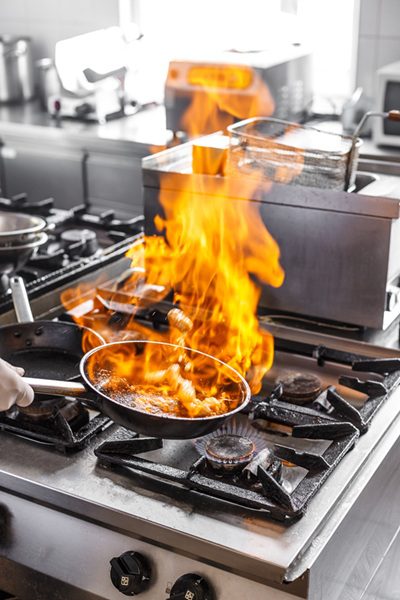 Flame in a frying pan in a busy restaurant kitchen, representing the risk of burn injuries for food service workers.