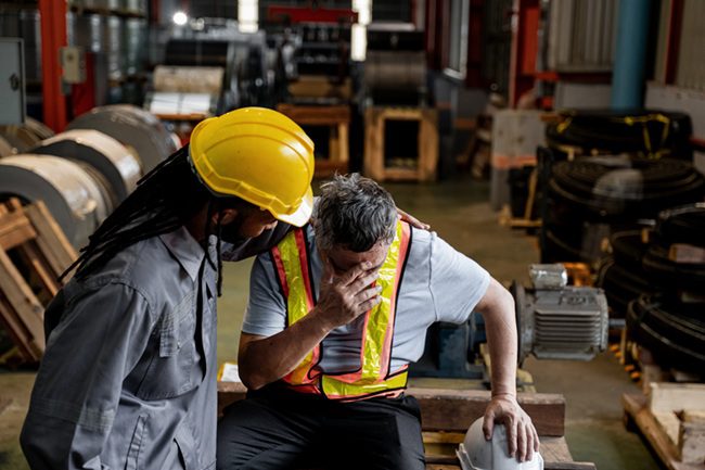 Two men, one wearing a yellow helmet and the other in a grey shirt, are sitting on a bench. The man in the yellow helmet is comforting the man in the grey shirt