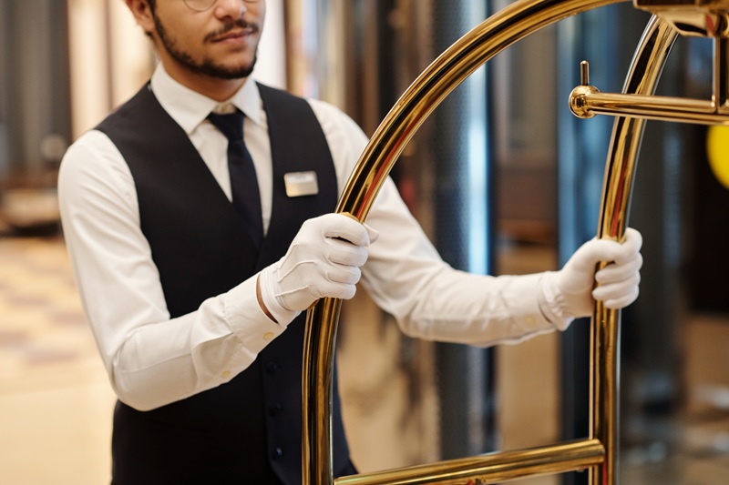 Close-up of elegant bellboy in uniform and white gloves pushing cart with baggage of tourists while moving along lobby in front of camera